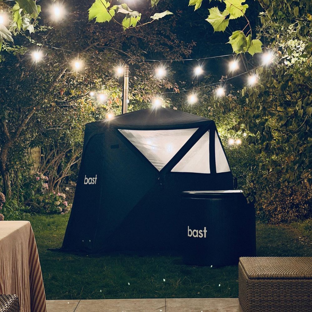 A black Bast sauna tent with white branding is pitched outdoors in a UK garden at night, lit by festoon lights.