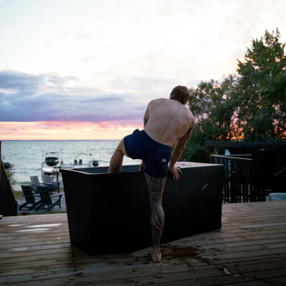 Man climbing into a cold plunge tub on a deck with a sunset over water in the background